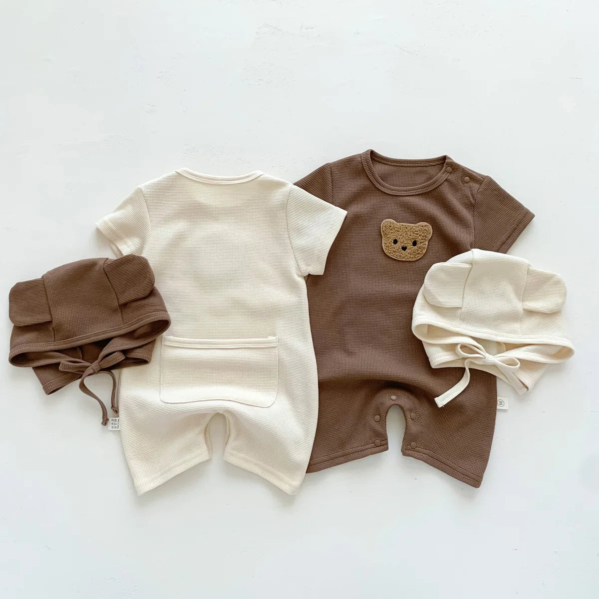 Two baby rompers, one cream and one brown with a bear face, are paired with matching bear-eared hats in cream and brown, arranged on a light background.