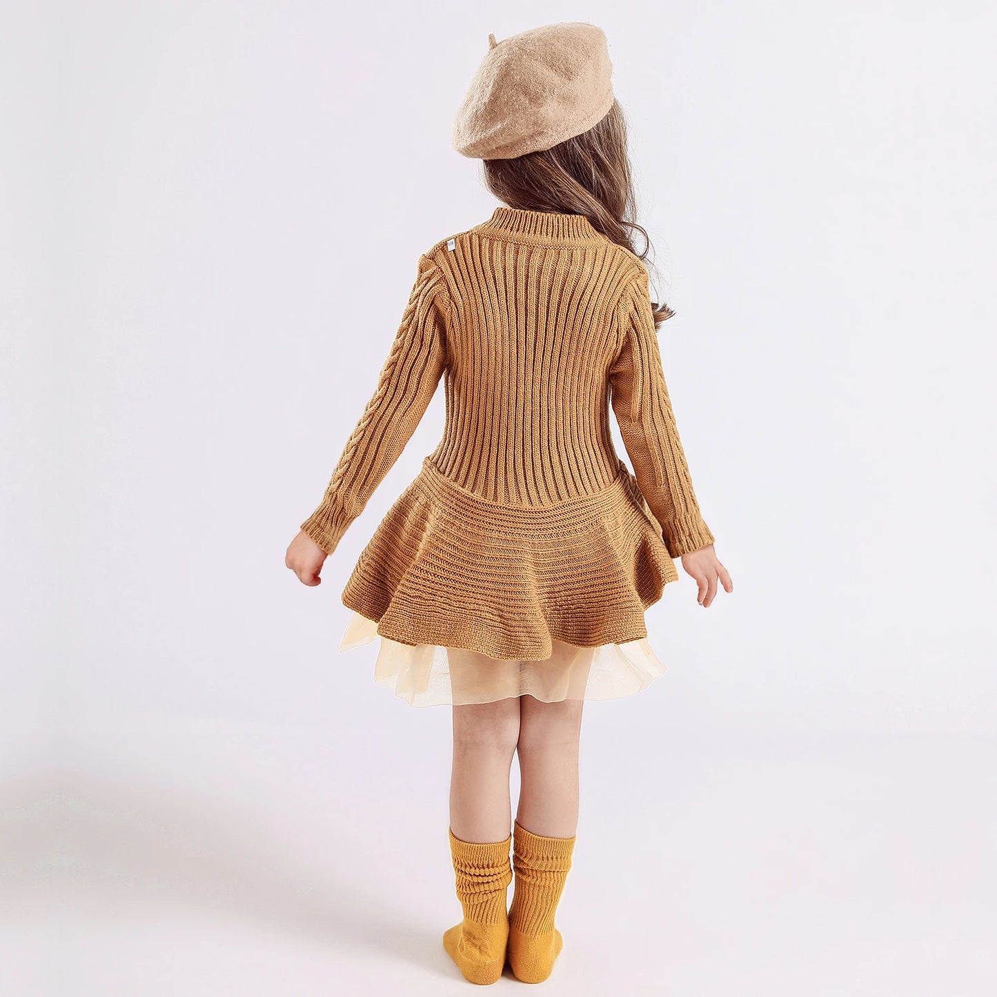 Young girl in a brown knit dress with a pleated skirt, matching beret, and yellow socks, facing away in a minimalist studio setting with a pale background.