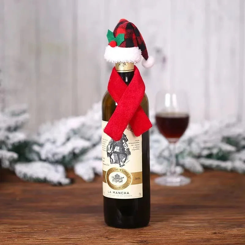 A wine bottle decorated with a red scarf and a small plaid Santa hat, set on a wooden table with frosted evergreen branches in the background. A glass of wine is partially visible.