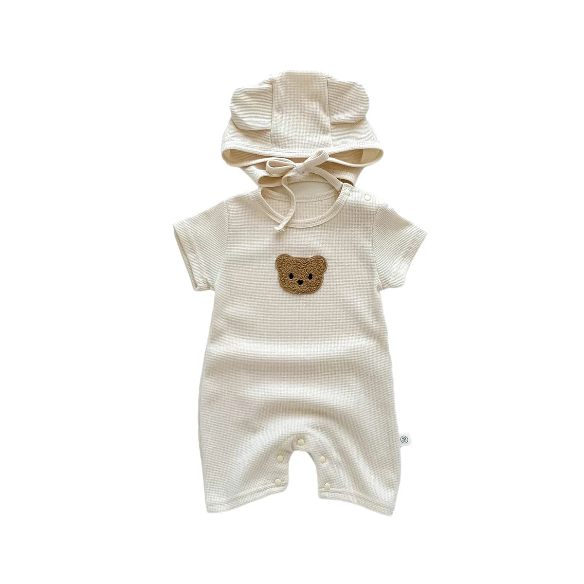 Cream baby romper with a teddy bear patch on the chest, featuring snap buttons on the legs and a matching bonnet with bear ears and a tied ribbon.
