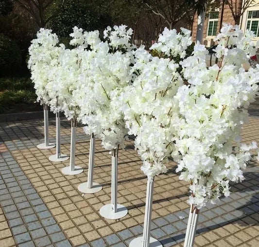 Rows of artificial white flower arrangements stand on tall white stands, positioned on a tiled outdoor surface, with trees and a building in the background.