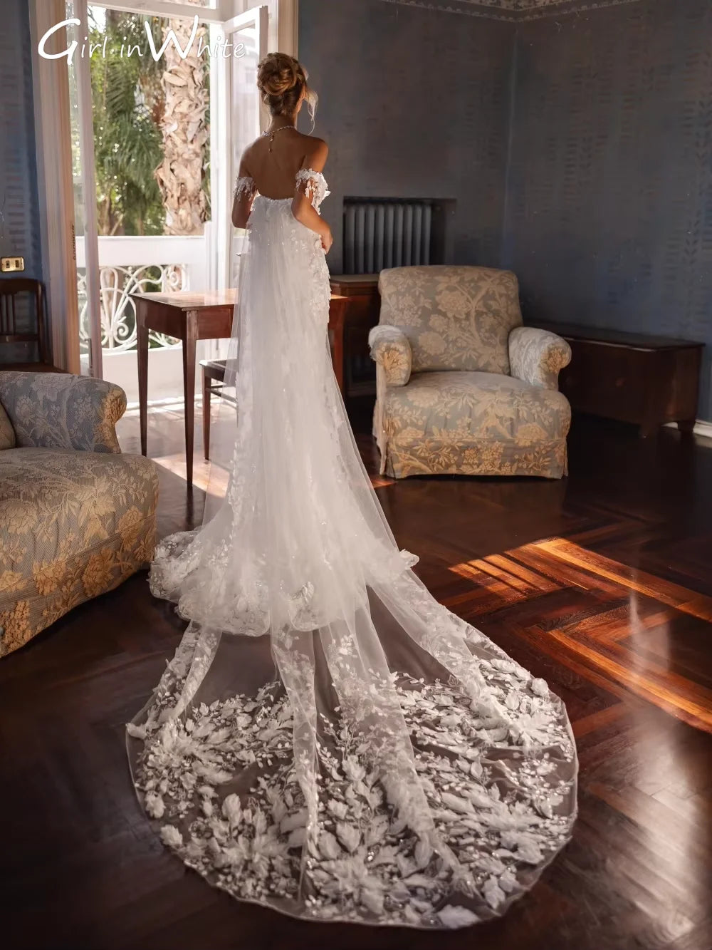 A woman in a long, flowing white lace wedding dress with an intricate train stands by an open door in an elegant room, with sunlight streaming in, highlighting her dress.