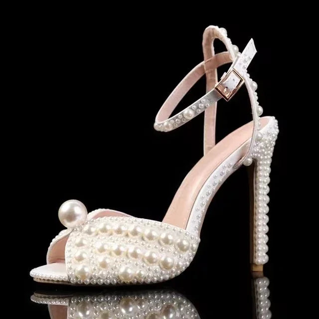 Elegant high-heeled sandal adorned with pearls, featuring an ankle strap and open toe. The heel is covered in pearls, set against a black background for contrast.
