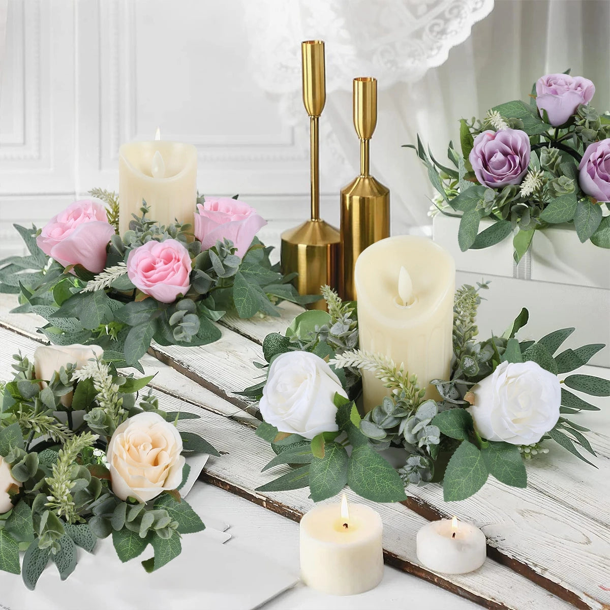 Elegant candles surrounded by pink, white, and lavender roses with greenery, placed on a rustic white wooden table. Two gold candle holders are in the background.