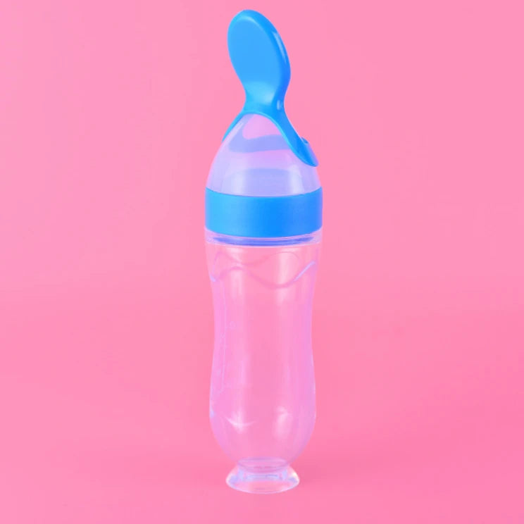A clear baby feeding bottle with a blue spoon-shaped lid, standing upright against a bright pink background. The bottle has a suction base for stability.