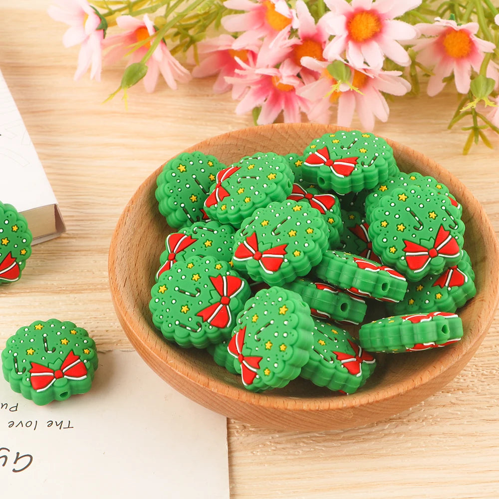 A bowl filled with decorative, tree-shaped charms featuring green foliage, red bows, and small yellow stars. Pink and white flowers are in the background on a wooden surface.