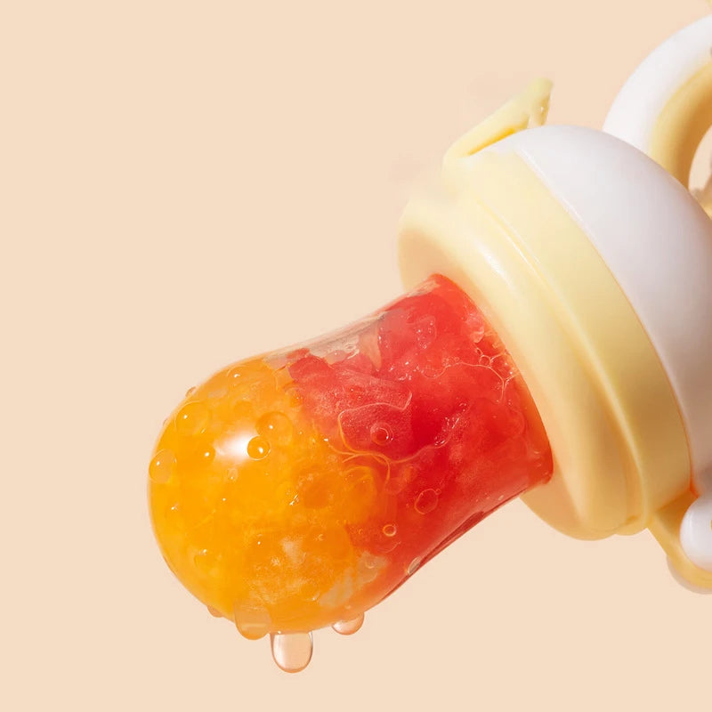 Close-up of a baby food feeder with a yellow and red fruit puree inside. The feeder has a yellow ring and a transparent silicone pouch against a peach background.