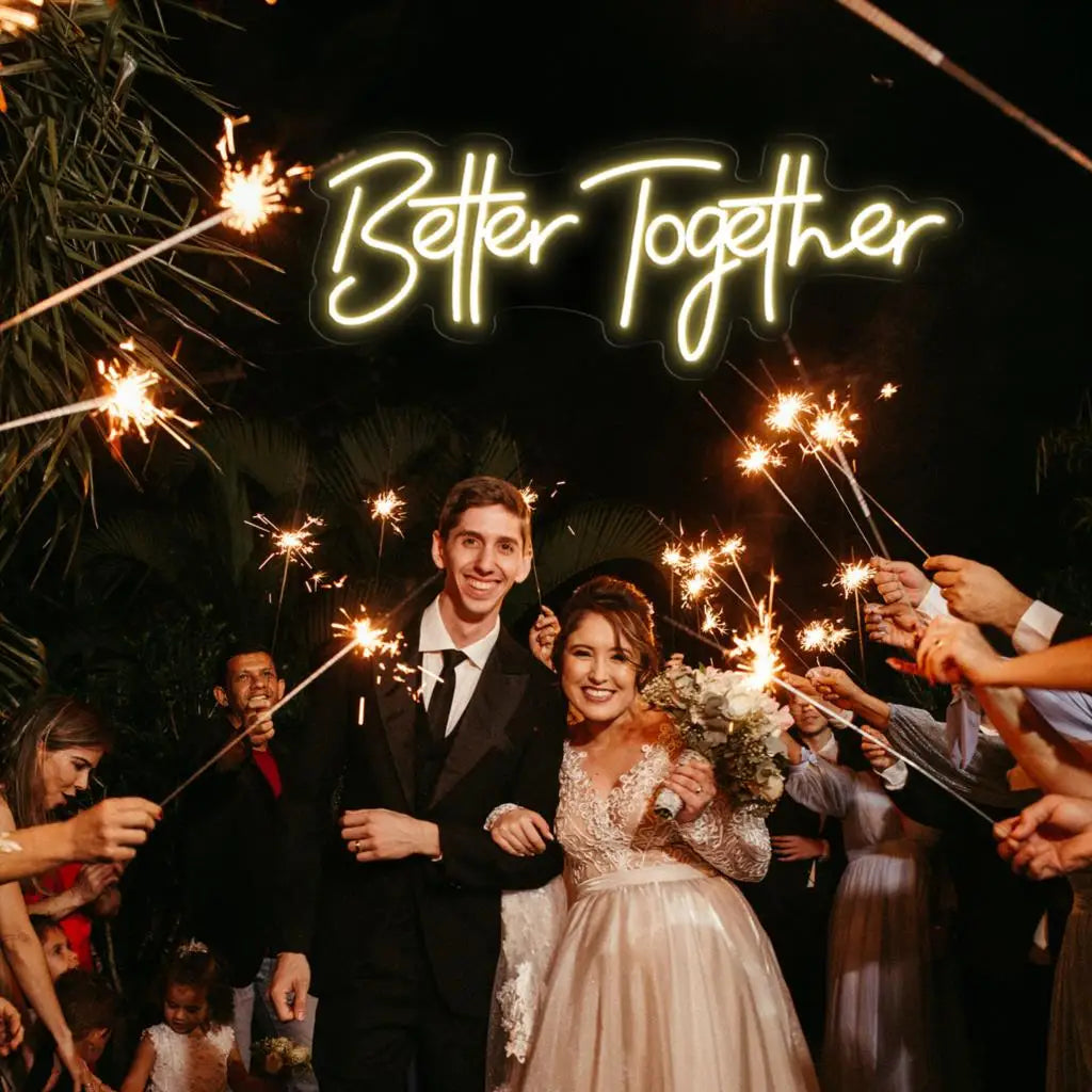 Bride and groom smiling, walking through a sparkler-lit path with guests. They are dressed in formal attire. Above them, a neon sign reads "Better Together."