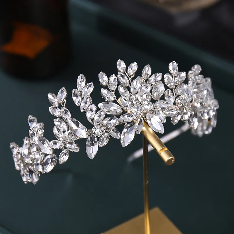 Sparkling tiara adorned with clusters of clear crystals set in a floral pattern, displayed on a golden stand against a dark background.