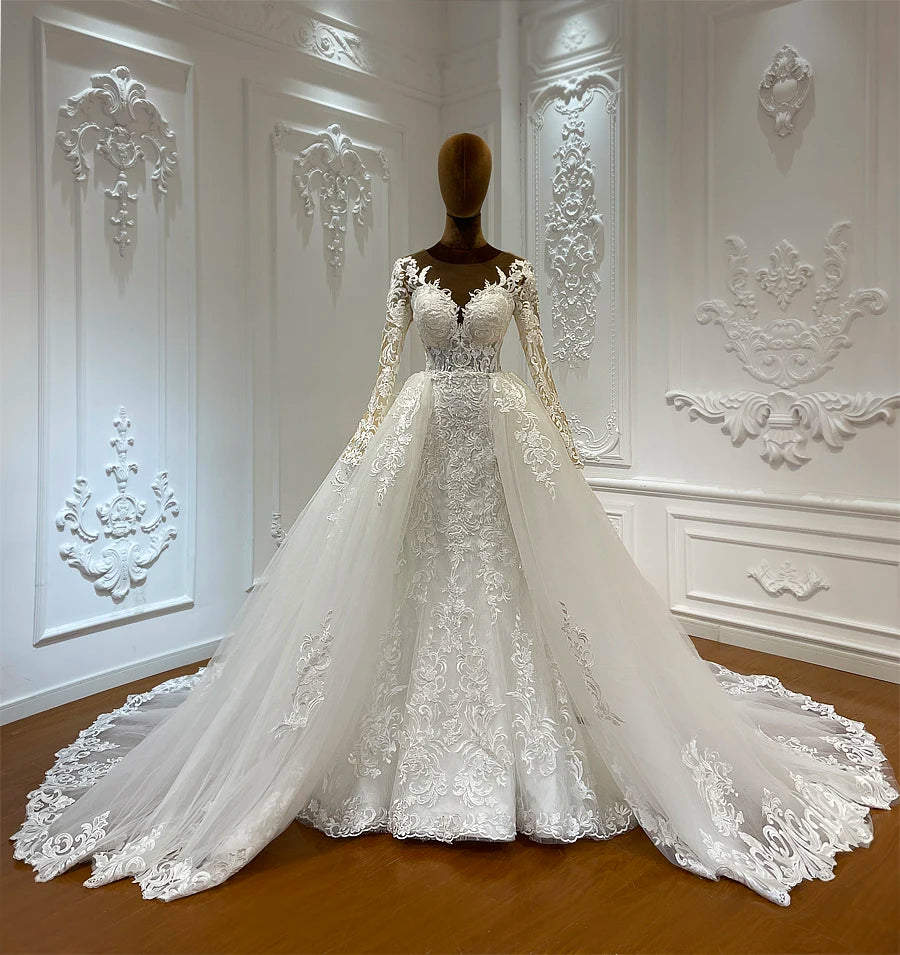 Elegant white wedding gown on a mannequin in an ornate room. The dress features long lace sleeves, an intricate bodice, and a voluminous layered skirt with lace details.