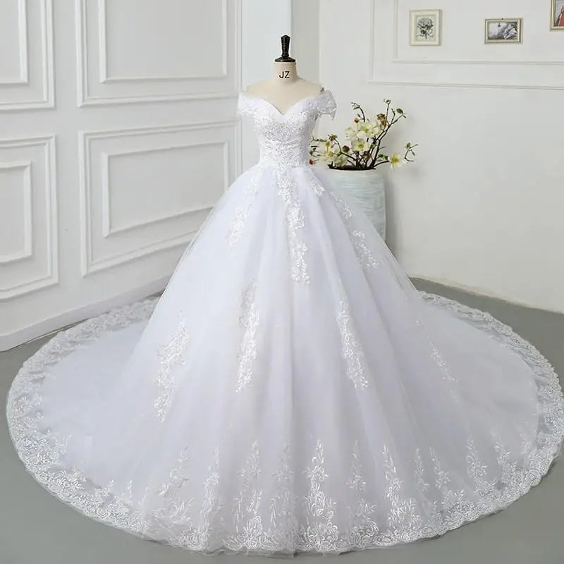 Elegant white wedding dress on a mannequin with an off-shoulder neckline, lace details, and a long flowing train. The backdrop features a minimalist, white-paneled wall and a vase with flowers.