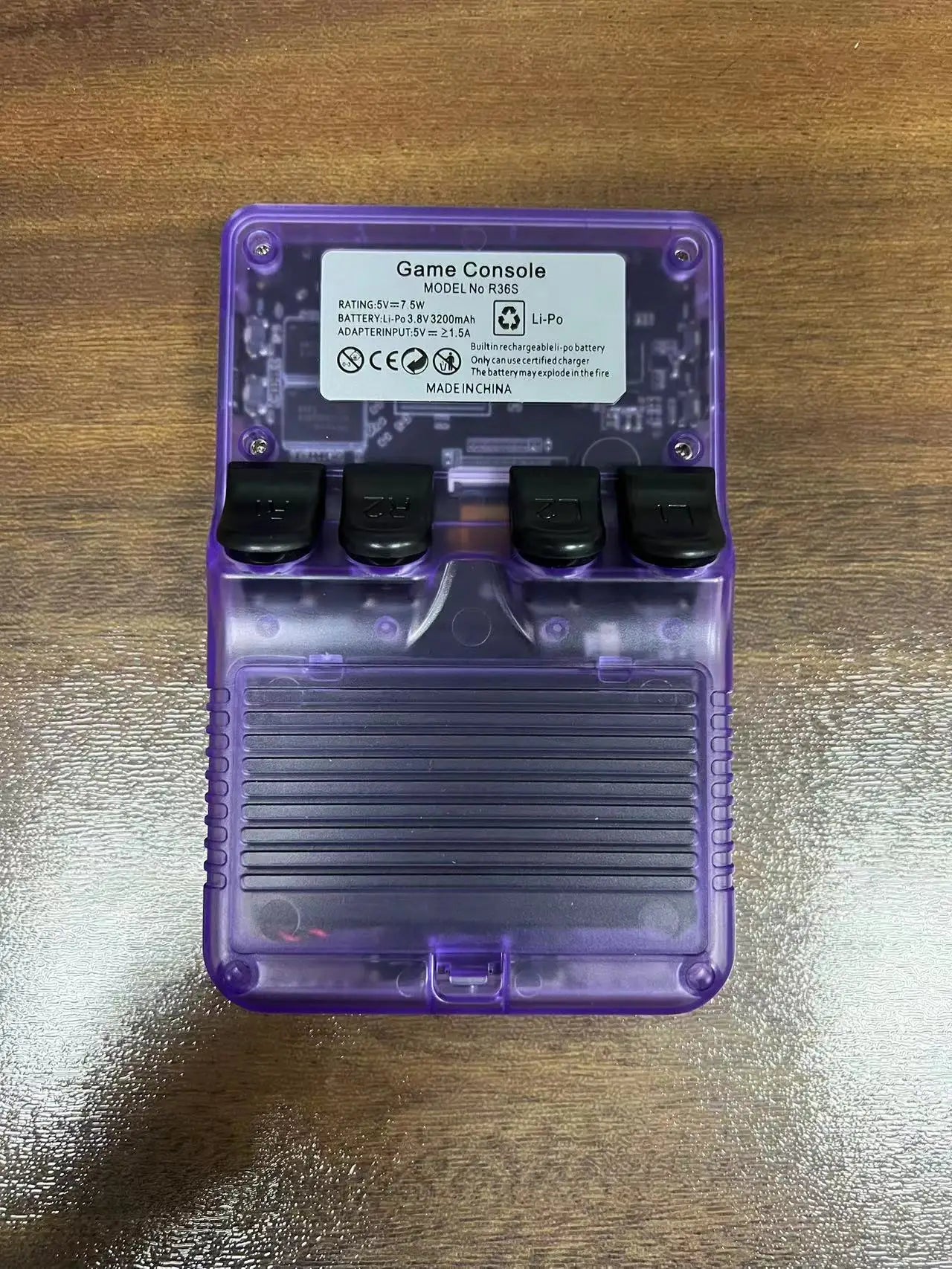 A purple, translucent R36S Retro Handheld Console – 64GB Games by Faron lies face-down on a wooden surface, showing its back cover, battery compartment, and label—a classic portable device for gaming enthusiasts.