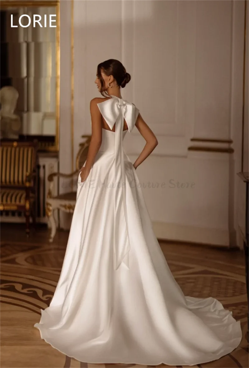 Elegant woman in a sleek, floor-length white gown with a large bow at the back, stands in a luxurious room with classical decor and gold accents.
