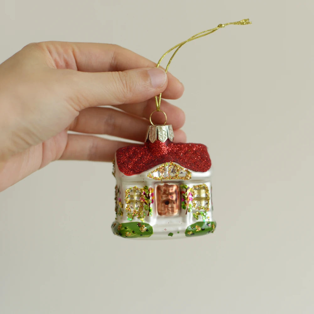Hand holding a festive house-shaped ornament with a red glitter roof, colorful decorations, and a gold string, against a plain background.