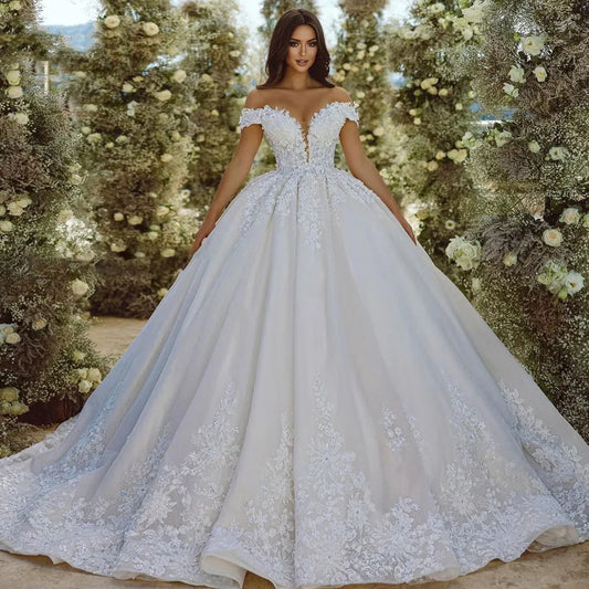 Bride in an elegant, off-shoulder white wedding gown with intricate lace details, standing in a lush floral archway adorned with white blossoms and greenery.