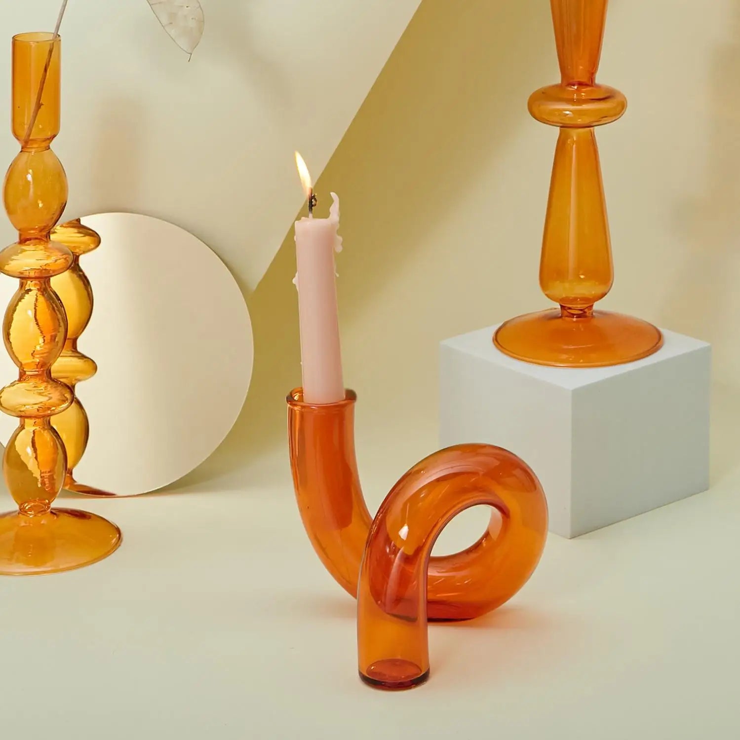 Curved orange glass candle holder with a lit white candle on a beige surface. Surrounded by two amber glass vases and a round mirror, creating a modern, minimalist aesthetic.