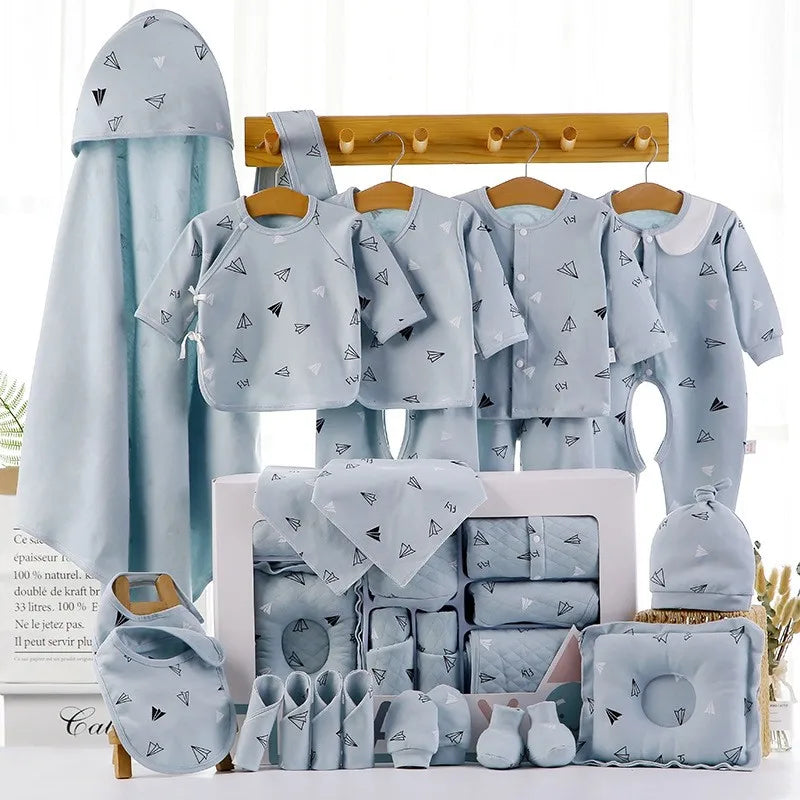 Set of baby clothes and accessories in light blue with paper airplane prints, including onesies, bibs, a hooded towel, mittens, and booties, neatly displayed on a rack and box.