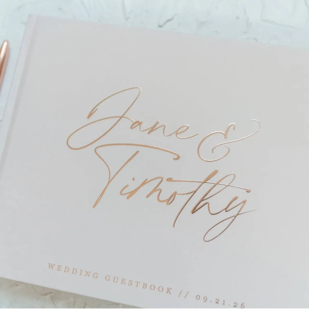 Elegant wedding guestbook with "Jane & Timothy" in gold script on a pale cover. Date "09.21.26" is subtly printed below. A pen is visible at the edge.