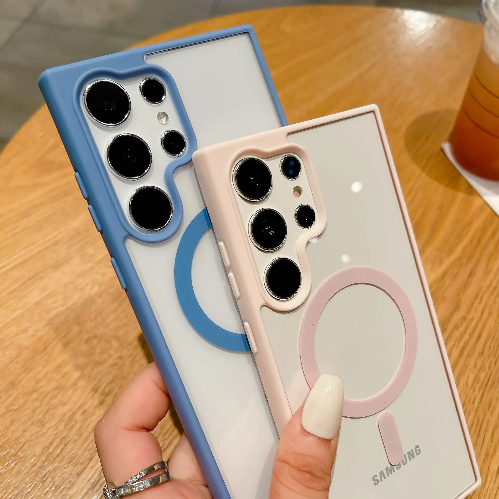 Two Samsung smartphones with protective cases; one blue, one pink. Each has a ring design around the camera lenses. They're held above a wooden table with a drink nearby.