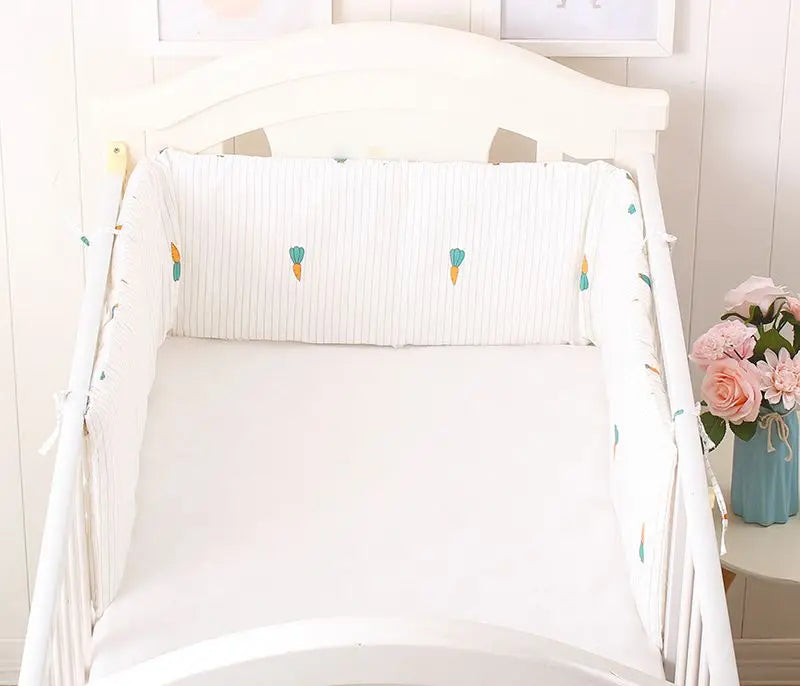 White crib with patterned bumpers featuring small balloons. A vase with pink flowers is on a side table. The room has a soft, pastel interior design.