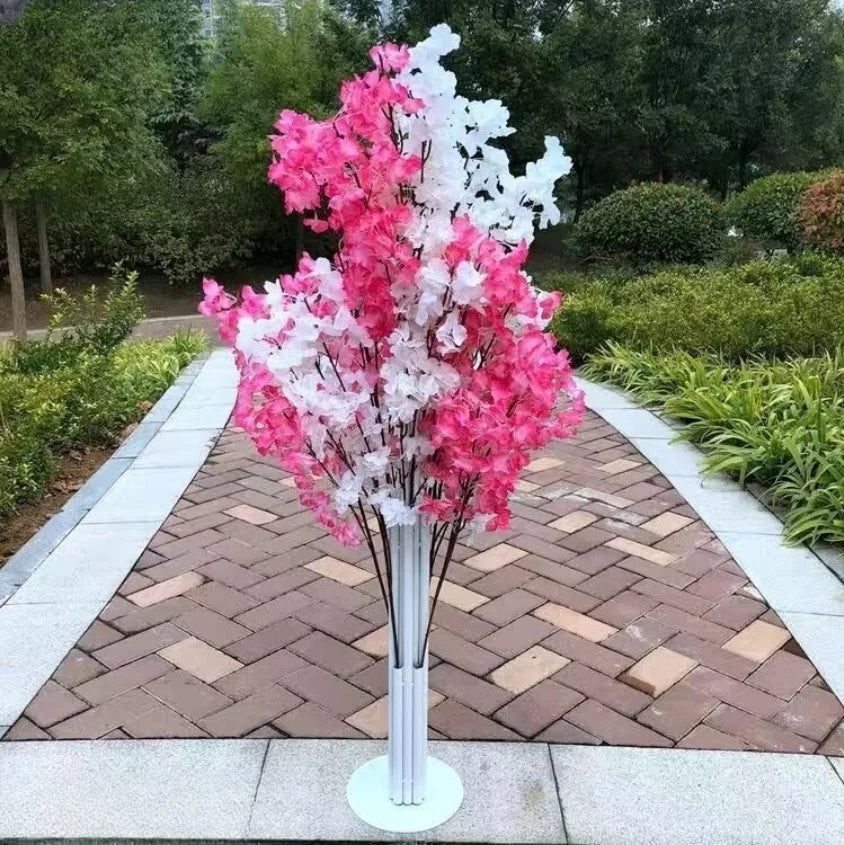 Pink and white artificial flowers in a tall white stand, placed on a paved pathway surrounded by green shrubbery and trees in a garden setting.