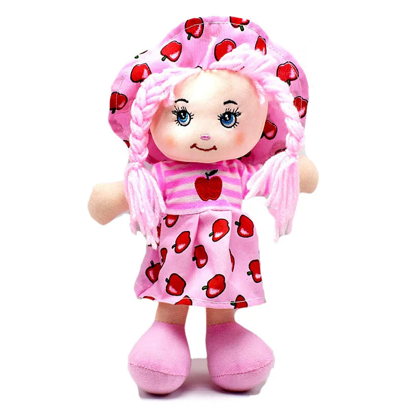 A plush doll with pink yarn hair, wearing a pink dress and hat with red apple prints. The doll has a friendly face and is also wearing matching pink shoes.
