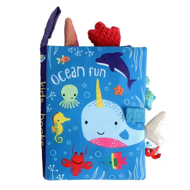 Colorful fabric book for kids titled "Ocean Fun," featuring a smiling narwhal, dolphin, octopus, seahorse, fish, crab, and vibrant sea-themed designs with soft toys attached.