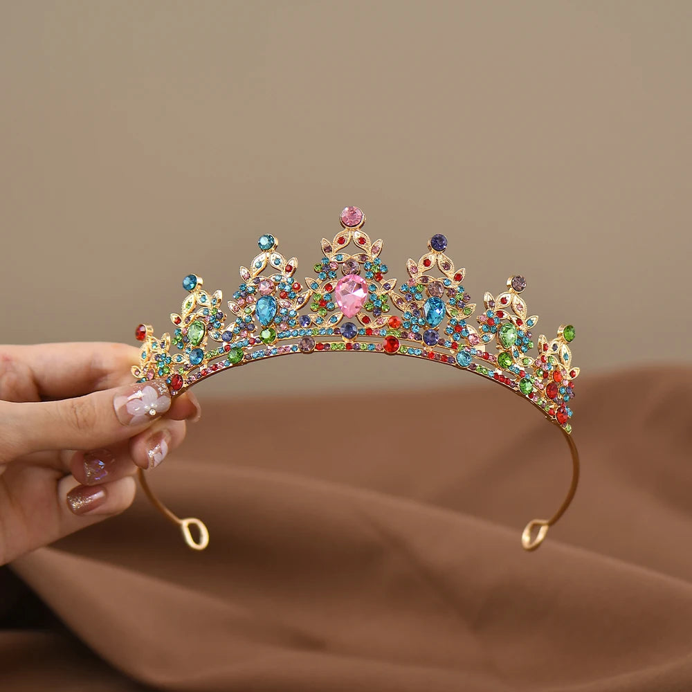 A hand holding an ornate gold tiara adorned with vibrant gemstones in various colors, including pink, blue, green, and red, set against a soft brown fabric background.