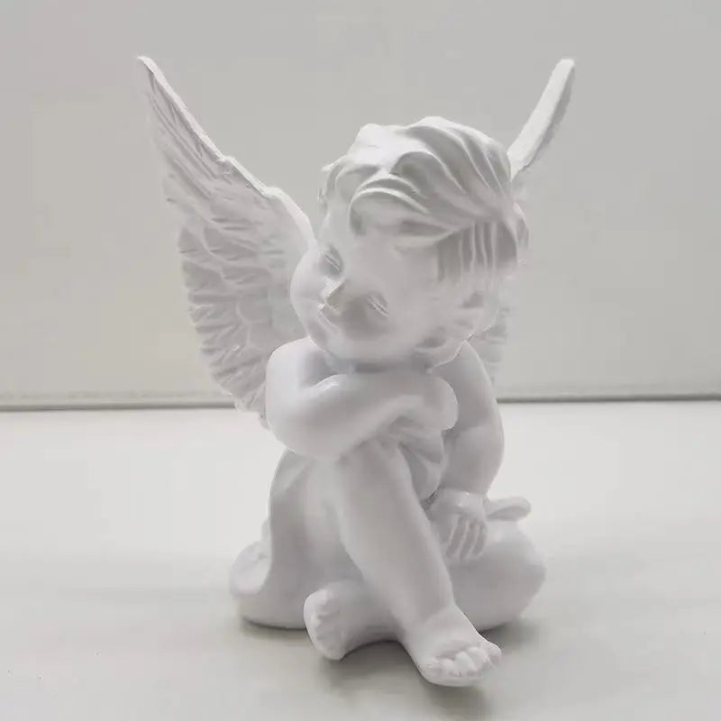 White porcelain cherub figurine with curled hair and feathered wings, sitting with one arm resting on its knee and the other around its leg, set against a plain background.