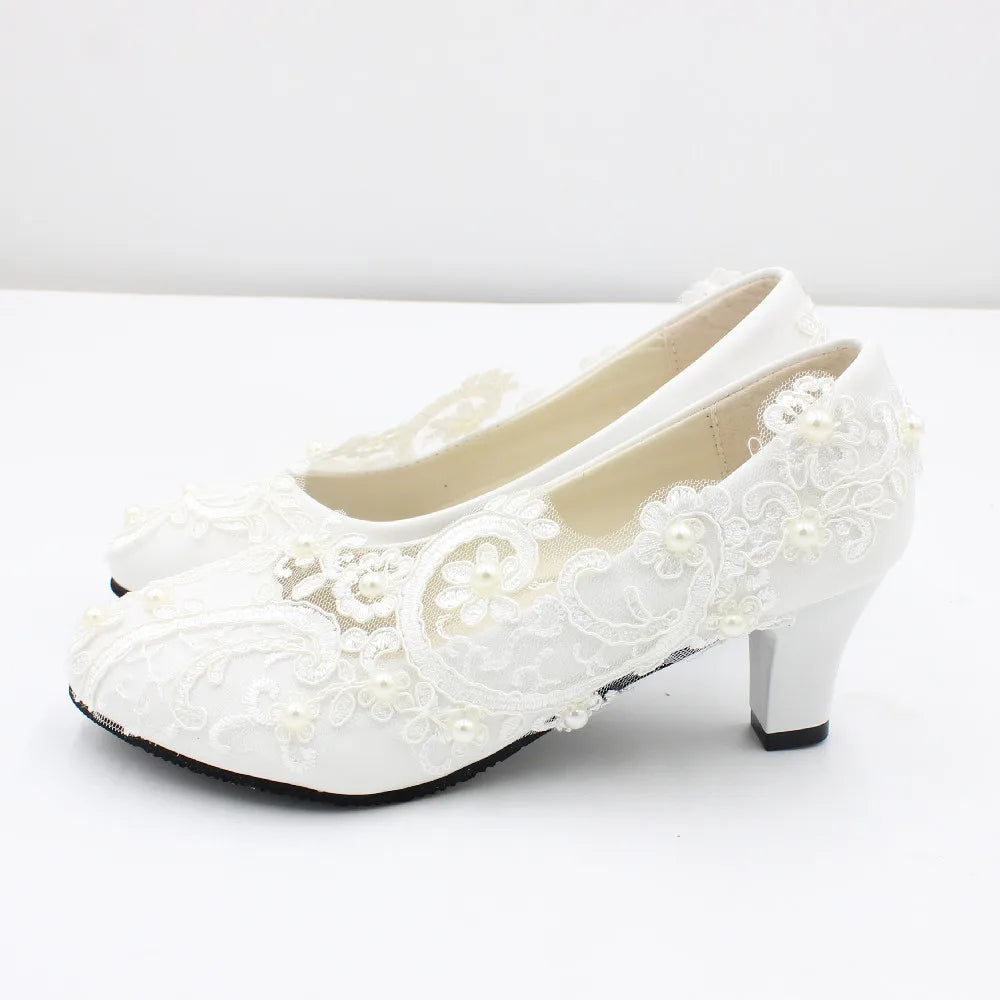 White high-heeled shoes adorned with intricate lace and small pearl-like embellishments, set against a plain background, showcasing elegant wedding or formal footwear.