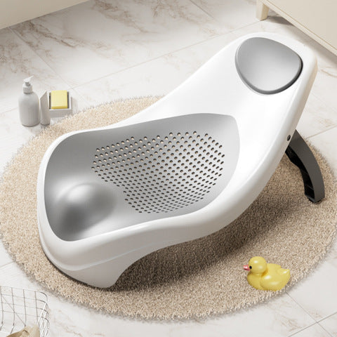 White and gray baby bathtub with perforated seat on a beige rug. Nearby are a small yellow rubber duck, a bottle, and a folded towel on a tiled bathroom floor.