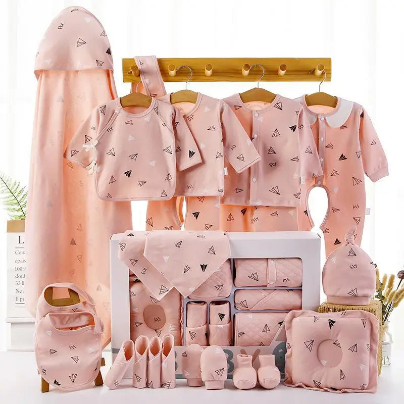 A collection of pink baby clothing and accessories with paper airplane patterns neatly arranged. Includes shirts, onesies, a bib, hat, socks, mittens, and a towel on a wooden rack.