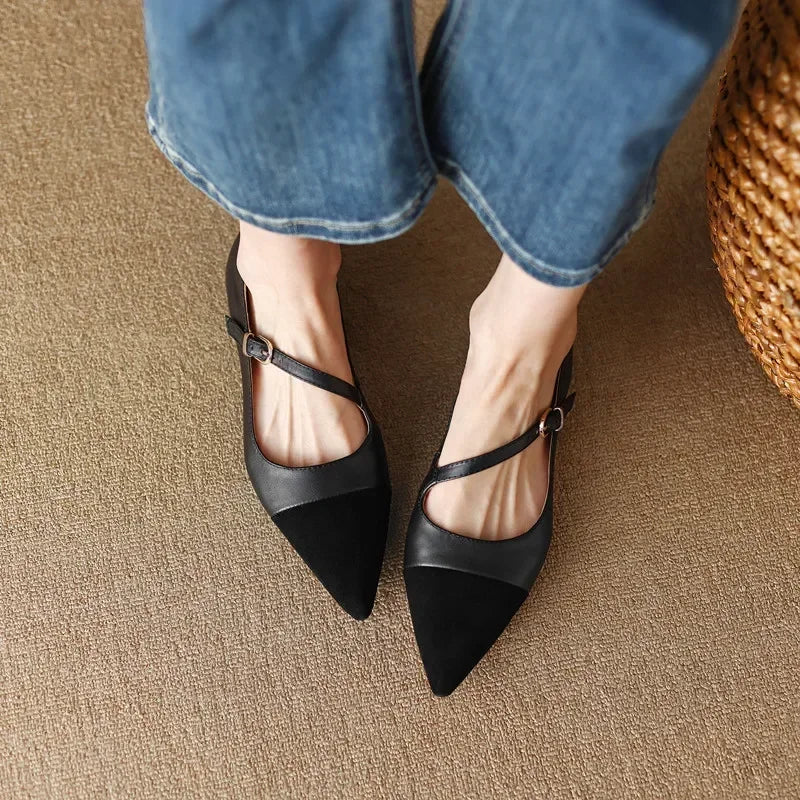 A person wearing black pointed-toe shoes with thin straps and buckles is standing on a beige carpet. Blue jeans slightly cropped at the ankles complete the casual look.
