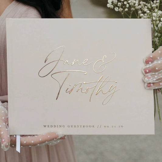 Wedding guestbook with "Jane & Timothy" in gold script held by a person in polka dot gloves, along with baby's breath flowers, dated 09.21.26.