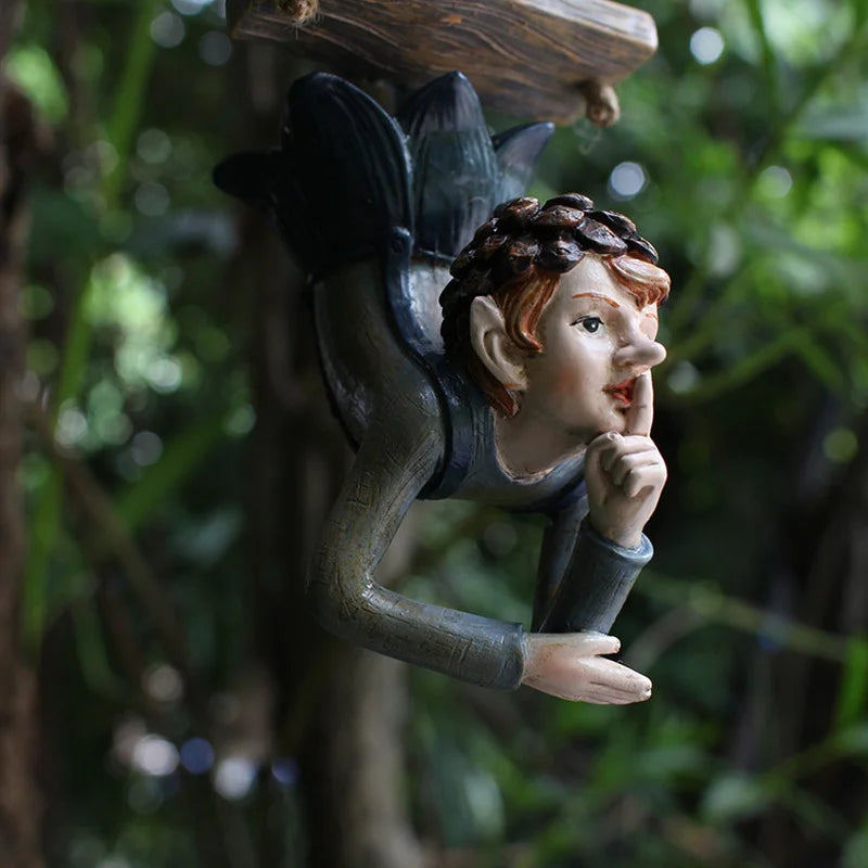 Whimsical garden sculpture of a fairy-like figure with pointed ears, placing a finger to its lips, suggesting silence. It hangs amidst lush green foliage, creating a mystical ambiance.