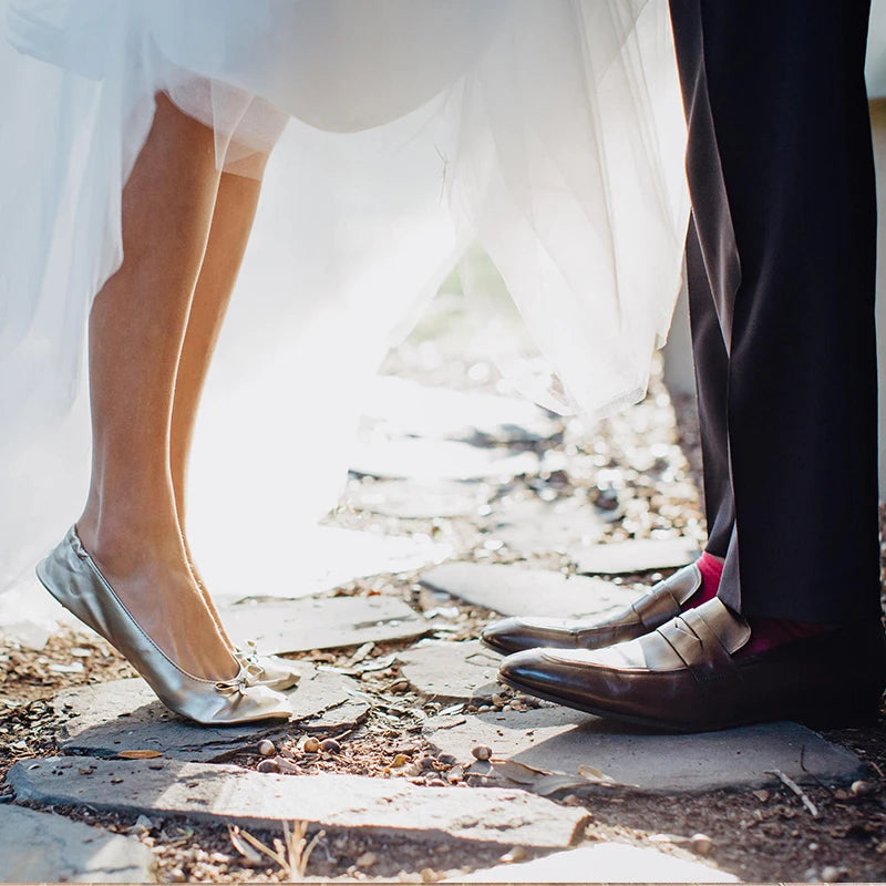 A bride in silver flats and a groom in formal shoes with red socks stand on a sunlit stone path, with her white dress flowing between them.