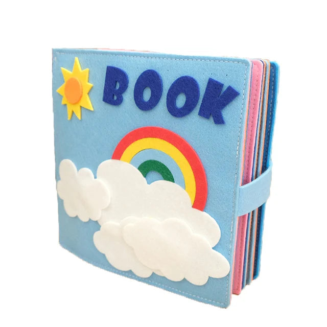 Colorful felt activity book with "BOOK" in blue letters, decorated with a sun, rainbow, and clouds on the cover. The book has a light blue cover and a buckle clasp.