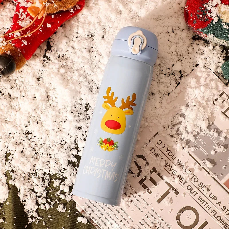 A light blue thermos with reindeer and "Merry Christmas" design lies on snowy ground beside a magazine. Red and green Christmas decorations are partially visible.