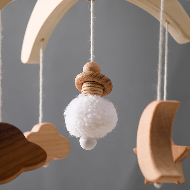 Wooden baby mobile with dangling shapes: a white fluffy pom-pom, clouds, and a crescent moon. All are suspended on strings against a neutral gray background.
