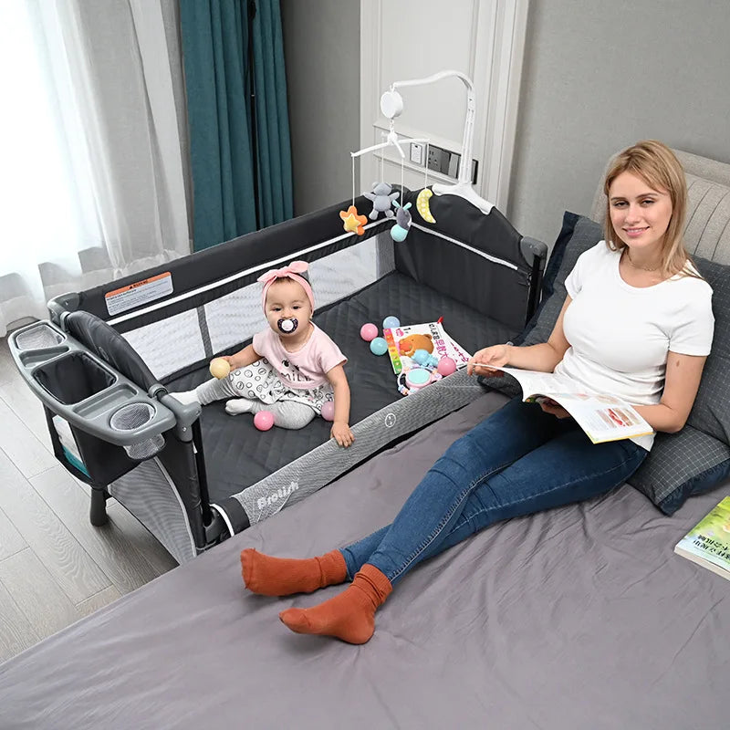 Woman relaxing with a book on a bed, next to a baby in a playpen. The baby has a pacifier and is surrounded by colorful toys, smiling under a mobile with hanging stars and moon.