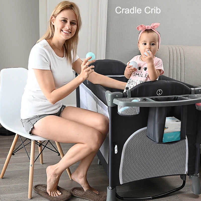 A woman in a white shirt sits in a chair beside a black cradle crib, interacting with a baby wearing a pink shirt and headband. The baby holds a bottle and toy.