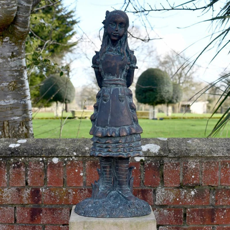 A bronze statue of a young girl with braided hair, wearing a ruffled dress and apron, standing on a pedestal. The background features trees and trimmed hedges in a garden setting.