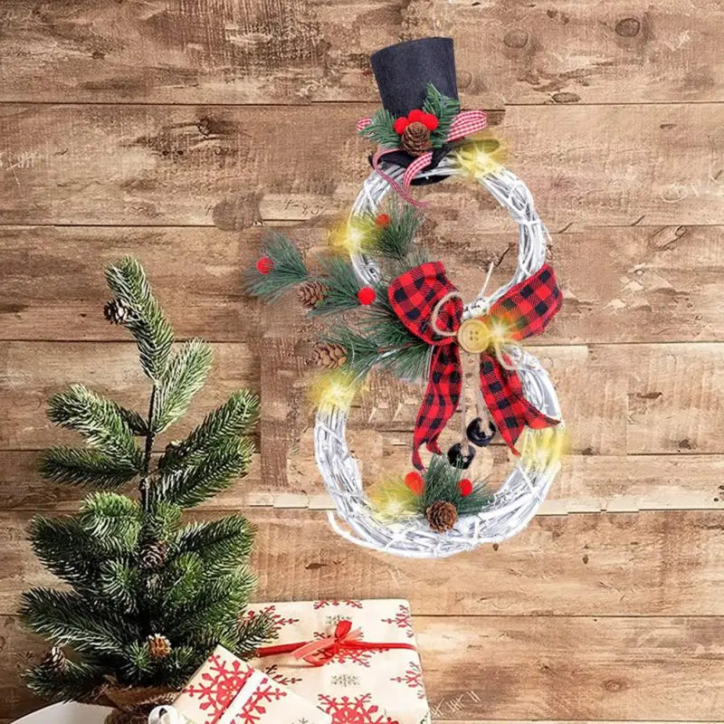 A festive snowman wreath with a top hat, plaid bow, pine cones, and red berries hangs on a wooden wall. A small pine tree and wrapped gifts are nearby.