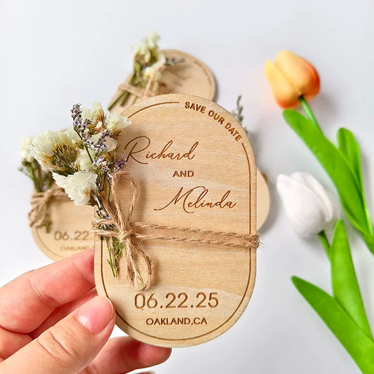 Wooden save the date card with floral decoration, reading "Richard and Melinda, 06.22.25, Oakland, CA." Held by a hand, with tulips in the background.