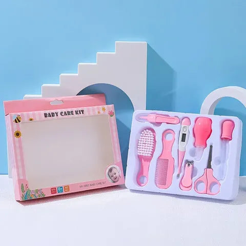 Pink baby care kit with a brush, comb, nail clippers, scissors, thermometer, and dropper. Set displayed against a pale blue backdrop next to an empty packaging box.