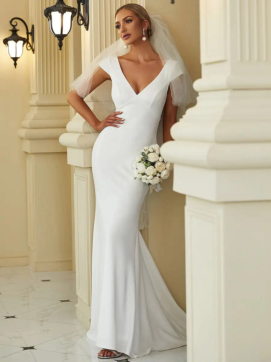 Bride in an elegant white gown with a deep V-neckline stands beside a column. She holds a bouquet of white flowers and wears a veil, with vintage wall sconces in the background.