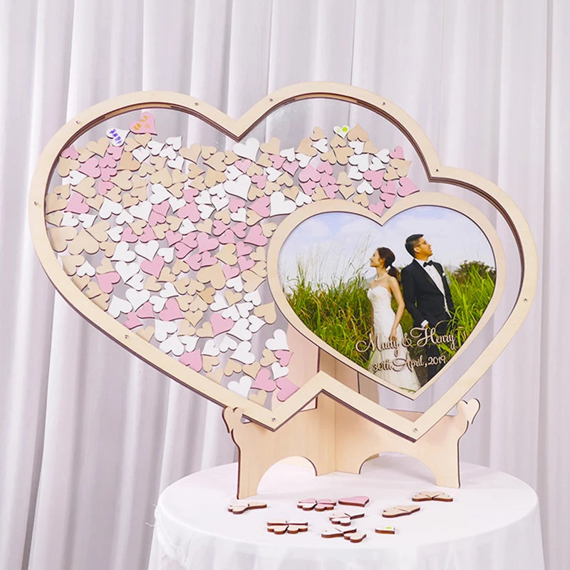 Heart-shaped wooden wedding guest book with pink and beige hearts. A photo of a couple is in the center. Displayed on a table with additional heart pieces scattered below.