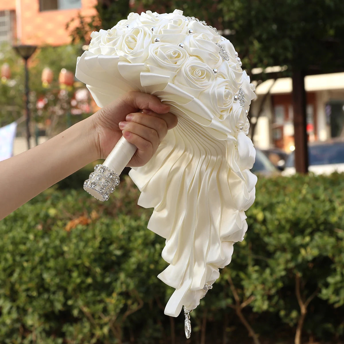 A hand holds an elegant bridal bouquet made of white satin roses with a jeweled handle. The background features greenery and blurred buildings.