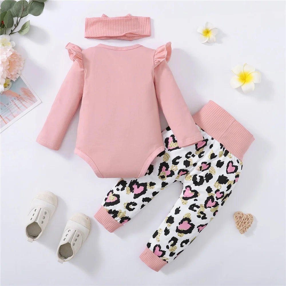 Pink long-sleeve onesie with ruffled shoulders, paired with leopard print pants in pink and black. Includes matching headband and ivory shoes, surrounded by flowers and decor.