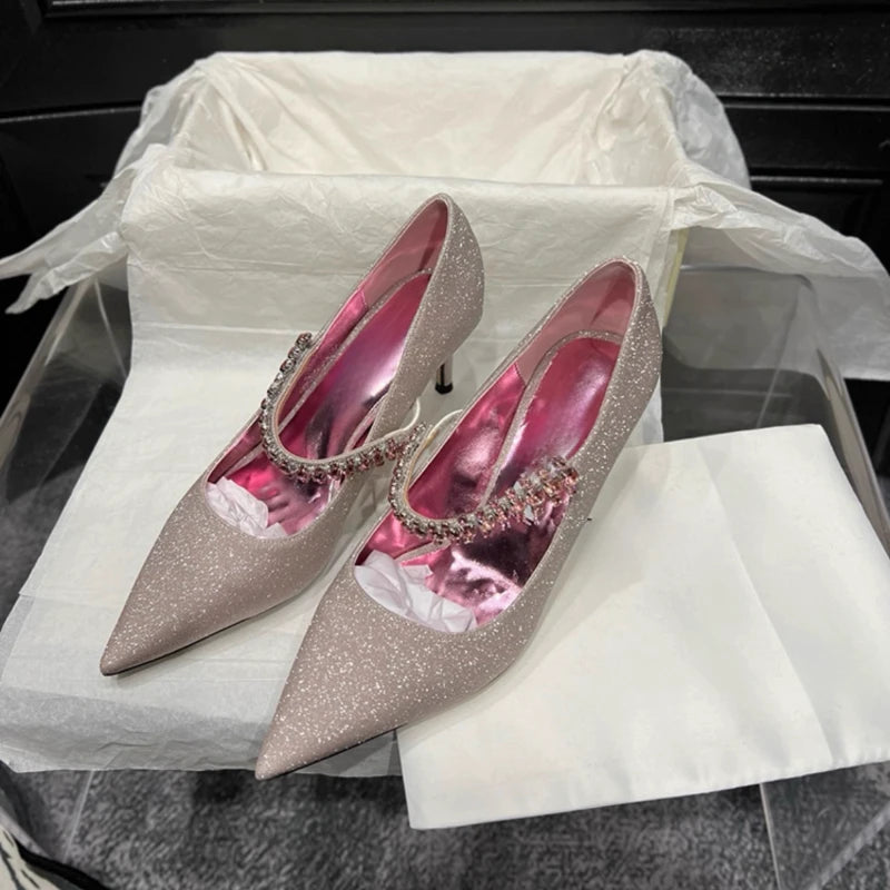 Sparkly beige high heels with pink insoles, decorated with a jeweled strap, displayed on white tissue paper in an open box on a black surface.